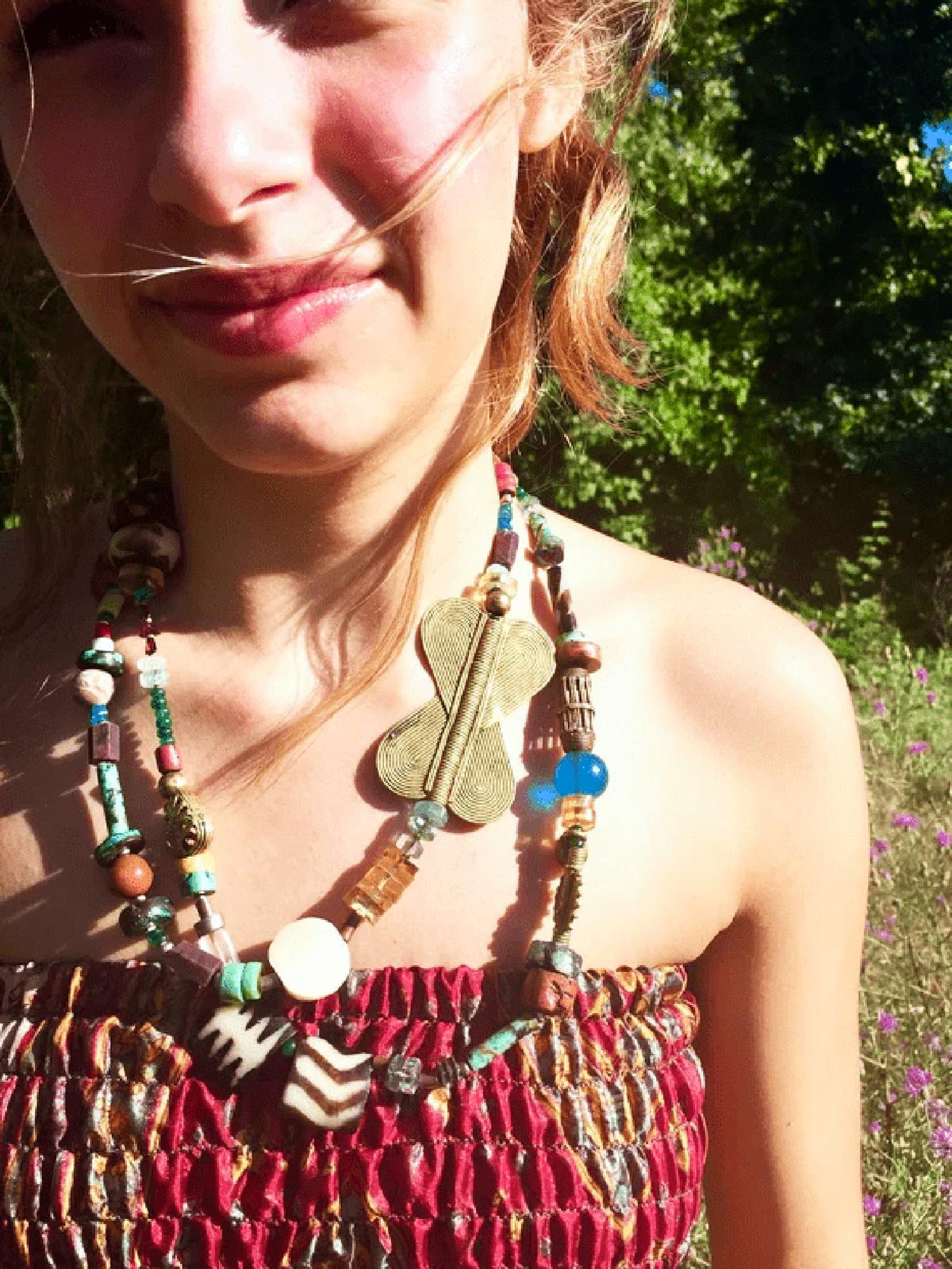 Outdoor portrait of a smiling young woman in sunlight, wearing a red smocked top and a handmade beaded necklace with eclectic, colorful beads and a prominent gold butterfly-shaped pendant. The background features green trees and purple wildflowers.
