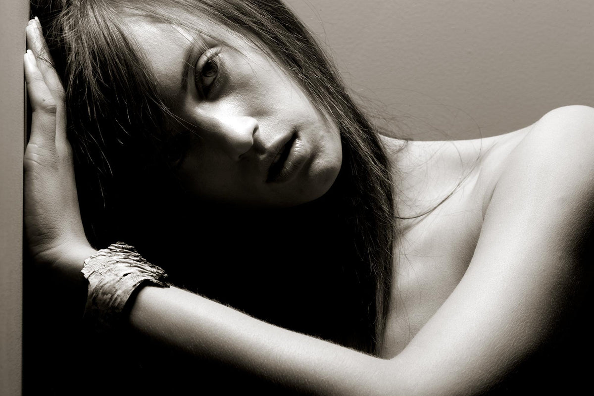 Black and white portrait of a woman leaning against a wall with her arms raised, wearing a textured bracelet and gazing directly at the camera.