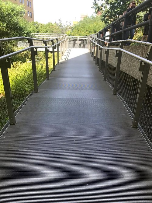 A wide, gently sloping ramp with metal handrails and mesh siding, surrounded by greenery and urban architecture in the background.