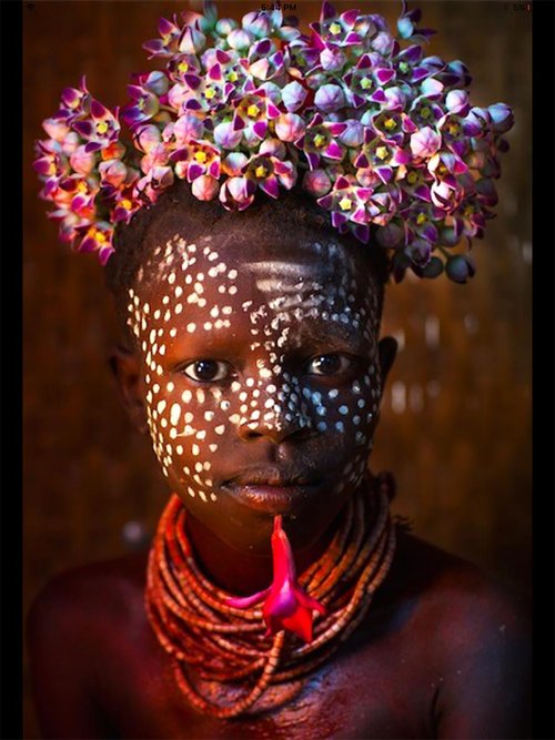 Portrait of a person with white face paint in dotted patterns, wearing a crown of pink and purple flowers and layered beaded necklaces, set against a warm-toned background.