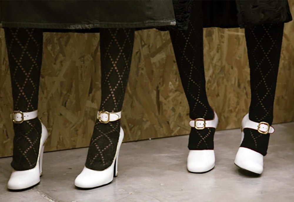Two pairs of legs wearing black diamond-patterned tights with white high-heeled Mary Jane shoes featuring gold buckles, standing against a plywood wall.