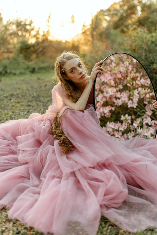 A young woman in a voluminous pink gown sits outdoors at golden hour, leaning on an oval mirror that reflects blooming pink flowers, creating a dreamlike and ethereal atmosphere.
