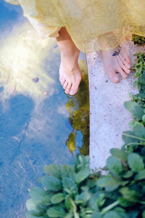 Bare feet with a small tattoo step delicately onto the edge of a stone ledge beside clear, shallow water, framed by green aquatic plants and the hem of a pale yellow dress.