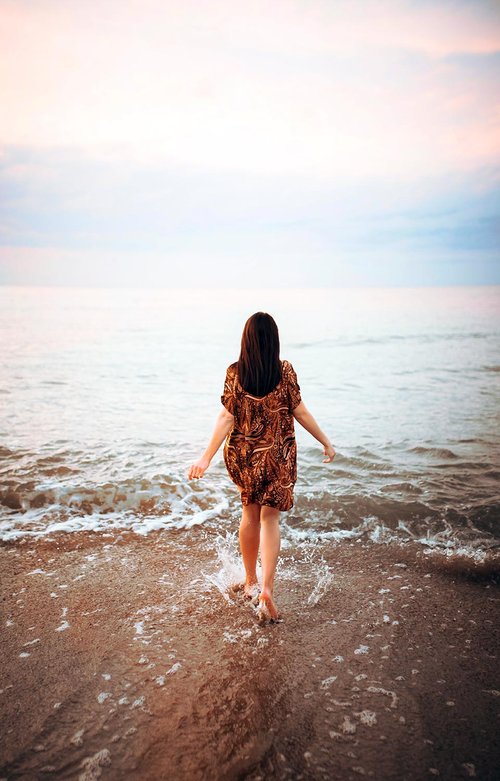 Woman in a patterned dress walking barefoot into the ocean during a soft pastel sunset, with gentle waves washing over the shore.