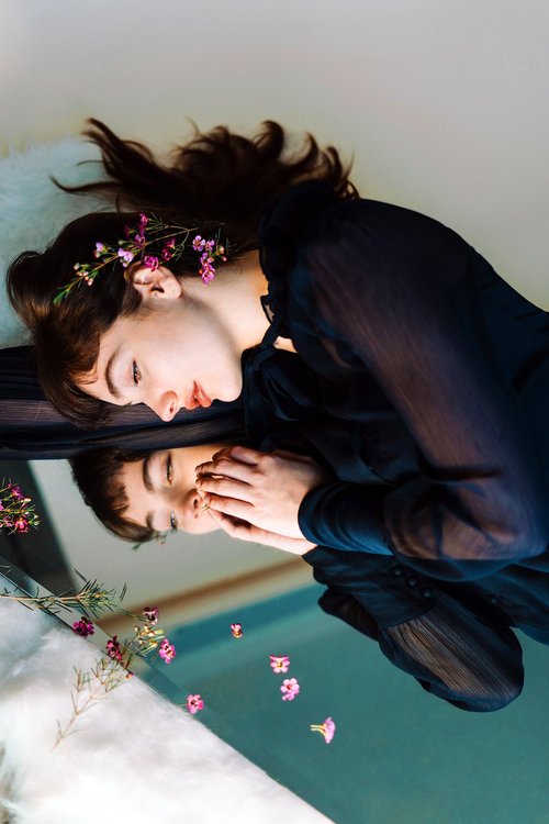 Woman in a sheer black dress lying beside a mirror with her hands gently touching her reflection, adorned with small pink flowers in her hair and scattered nearby.