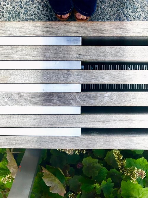 Top-down view of a slatted wooden bench with metal accents, a person’s feet in sandals above it, and green plants with small flowers below.