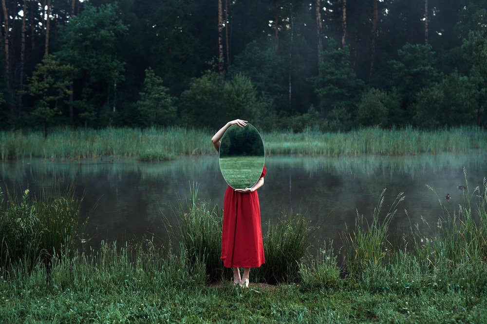 Person in red dress holding a mirror in front of a misty lake, reflecting only the grass behind them.