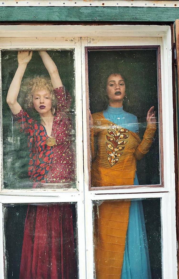 Two women standing behind a rustic window, wearing bold, vintage-inspired outfits—one in a red patterned dress with a large circular pendant necklace, and the other in a mustard and blue dress with intricate embroidered detailing, both showcasing elegant handmade jewelry in a moody, artistic setting.