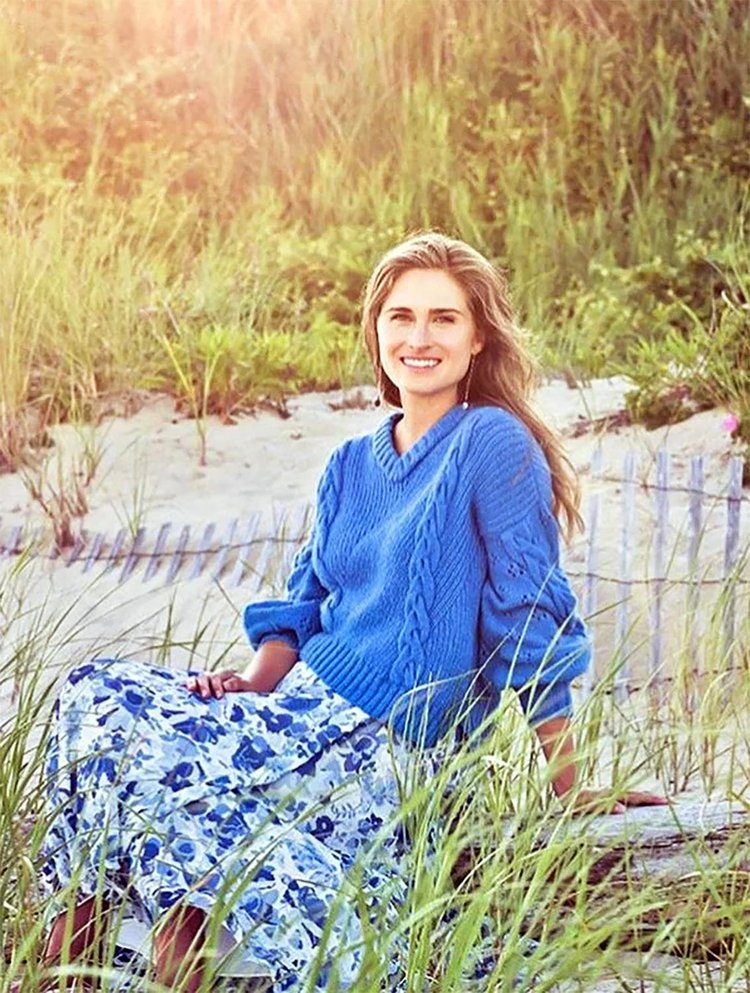 Woman sitting on driftwood at the beach surrounded by tall grass, wearing a bright blue cable-knit sweater and a flowing floral skirt, smiling warmly and accessorized with delicate handmade earrings for a breezy, sunlit coastal look.