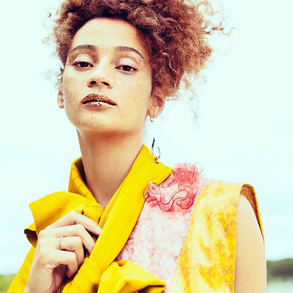 Close-up of a model with natural curls, gold glitter lips, and freckles, wearing a vibrant yellow and pink textured outfit adorned with ruffled fabric accents, and a dangling handmade earring that adds a bold, artistic flair to the editorial look.