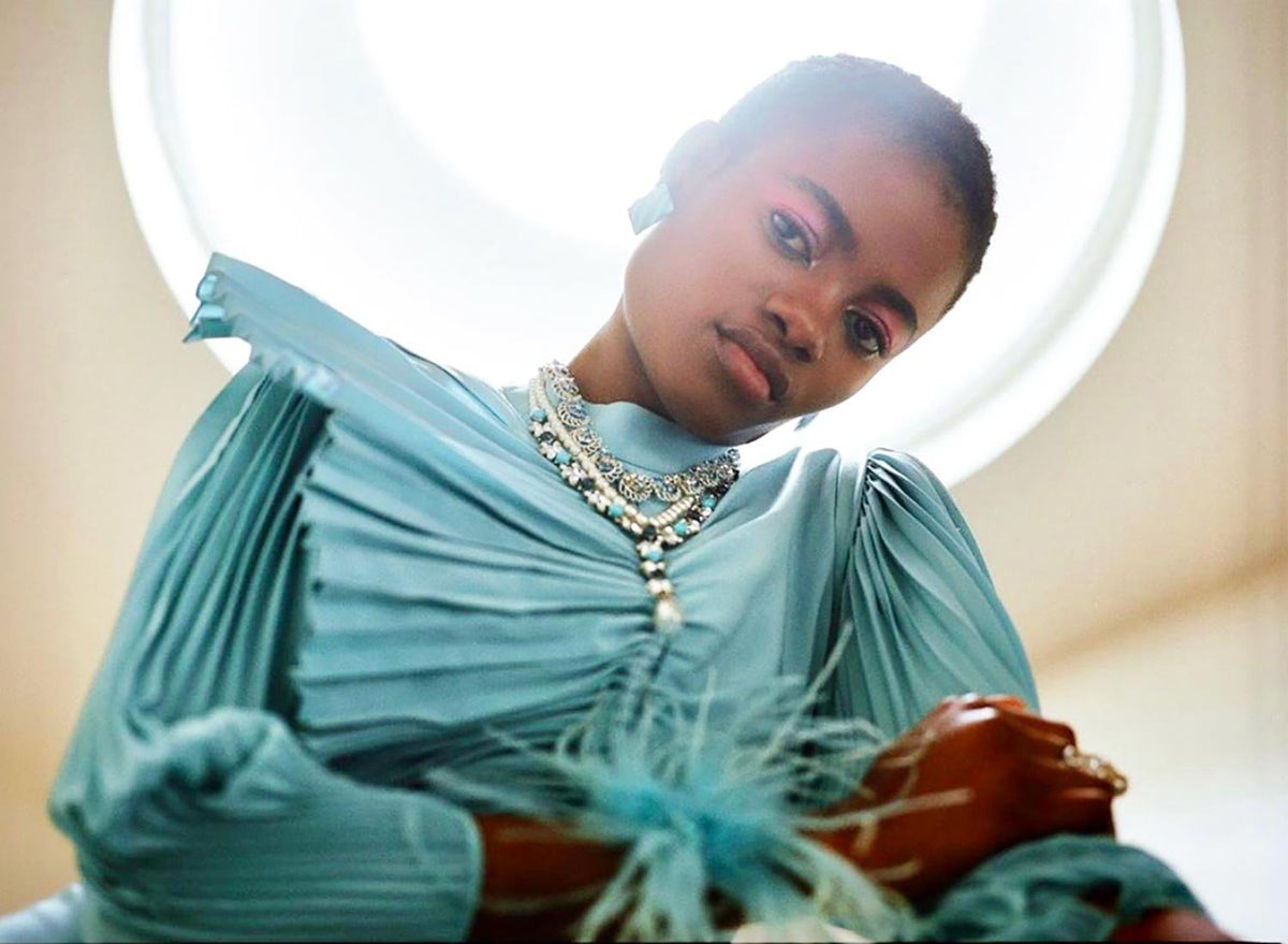 Model in a pleated, pastel blue gown adorned with feathered accents, wearing layered handmade necklaces with shimmering silver and turquoise details, posed beneath a circular skylight that casts a radiant glow around her elegant, editorial look.