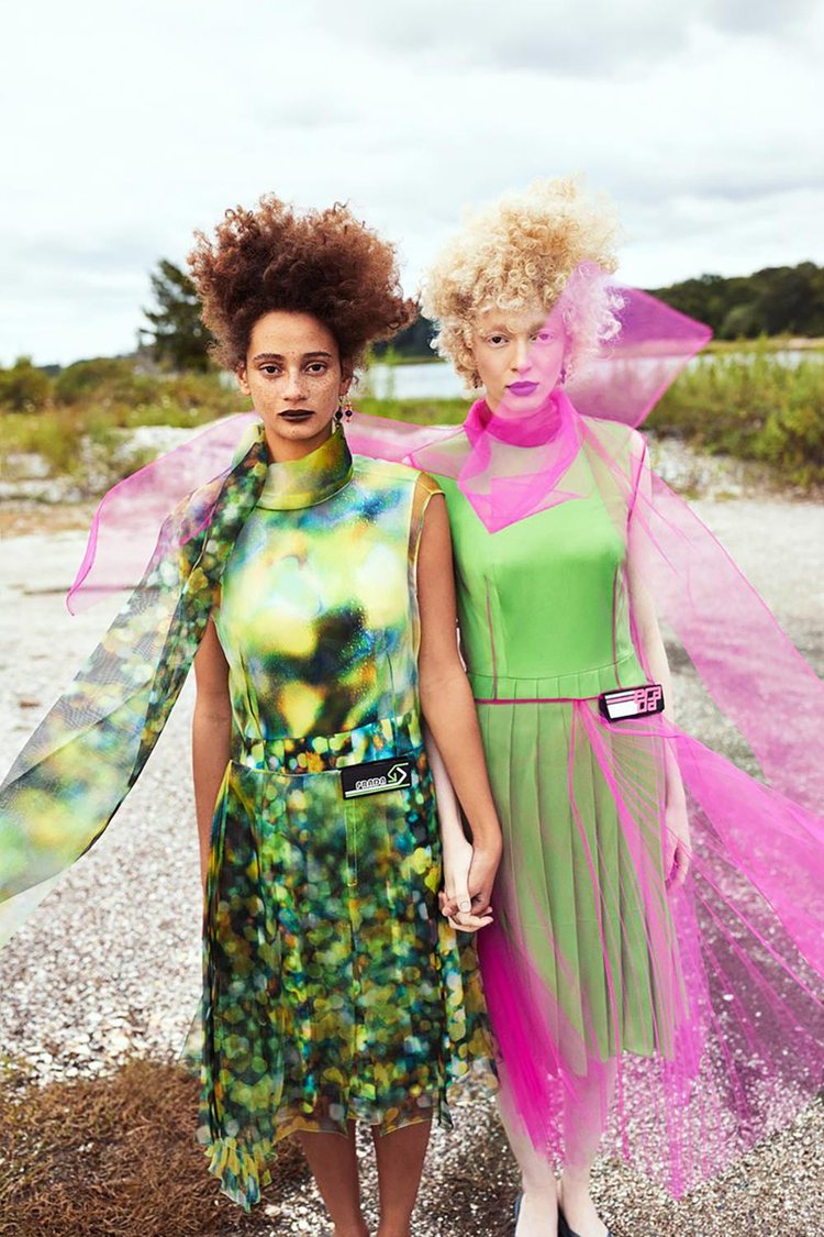 Two models standing hand in hand outdoors, dressed in futuristic dresses—one in a bokeh-inspired multicolor print and the other in neon green with sheer pink tulle—styled with bold lipstick, sculptural earrings, and expressive hair for a striking fashion-forward editorial.
