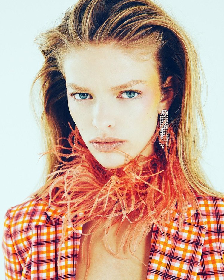 Close-up of a woman wearing a bold orange plaid blazer with a dramatic feathered collar and sparkling rhinestone chandelier earrings, showcasing a fierce, high-fashion look with handmade statement jewelry.
