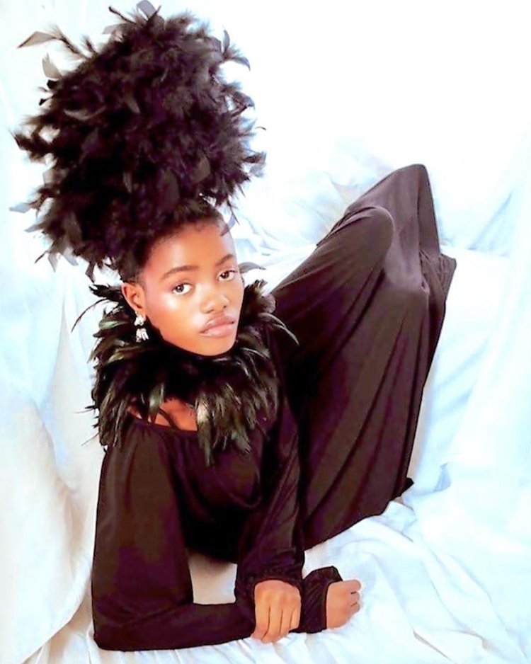Model posed against a soft white backdrop wearing a flowing black gown with dramatic feather detailing at the collar and an extravagant feathered headpiece, accessorized with elegant handmade earrings for a bold, regal editorial look.