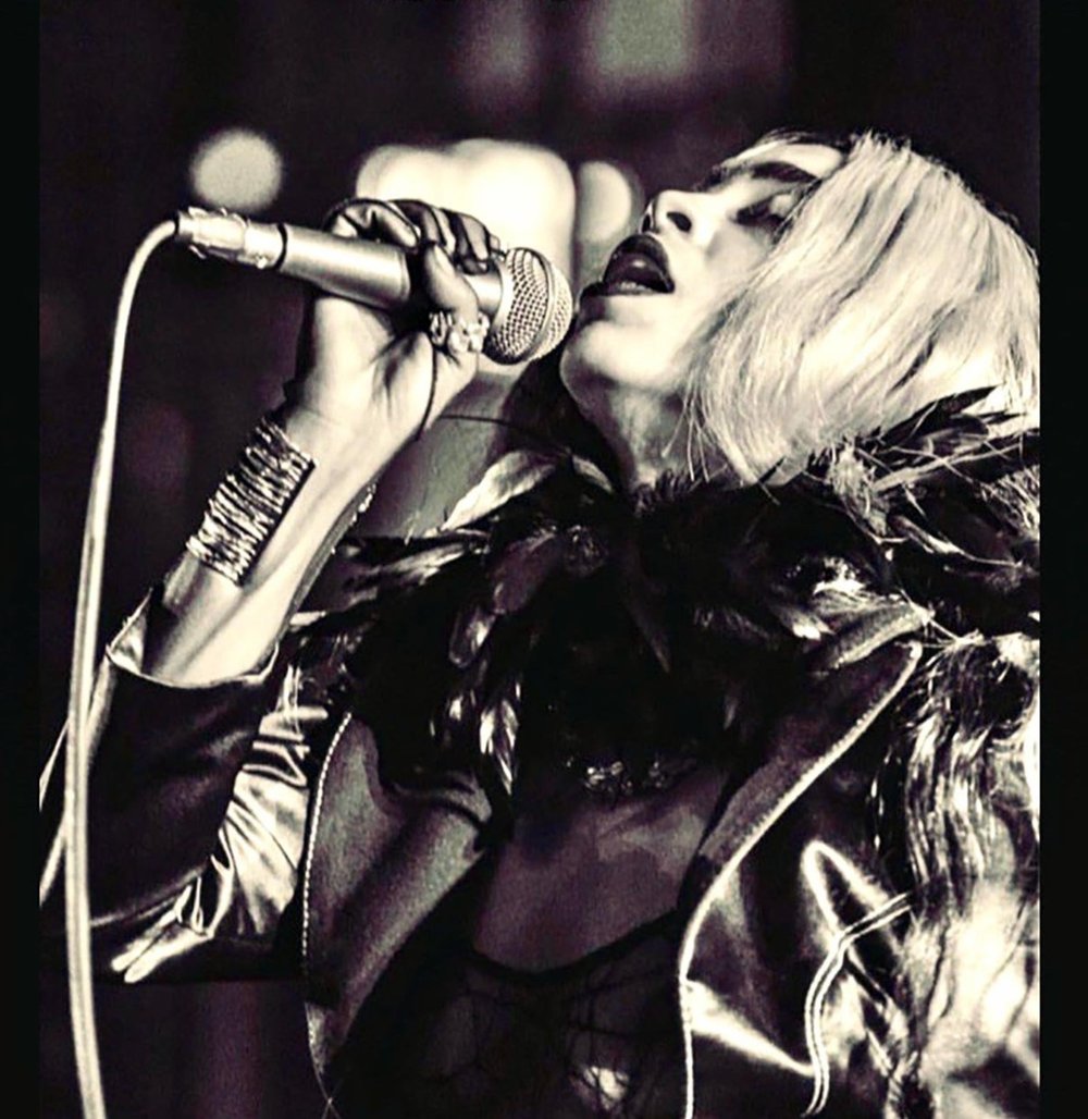 Dramatic black and white image of Kelsey Lu mid-performance, head tilted back in passion while holding a microphone, wearing a feathered collar, metallic jacket, and stacked handmade bangles that emphasize the bold, expressive energy of the moment.