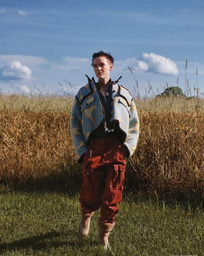Model walking through a grassy field under a blue sky, wearing a cropped wool jacket with abstract patterns, deep red cargo pants, fur-trimmed boots, and a chunky handmade necklace that adds a bold accent to the earthy, editorial look.