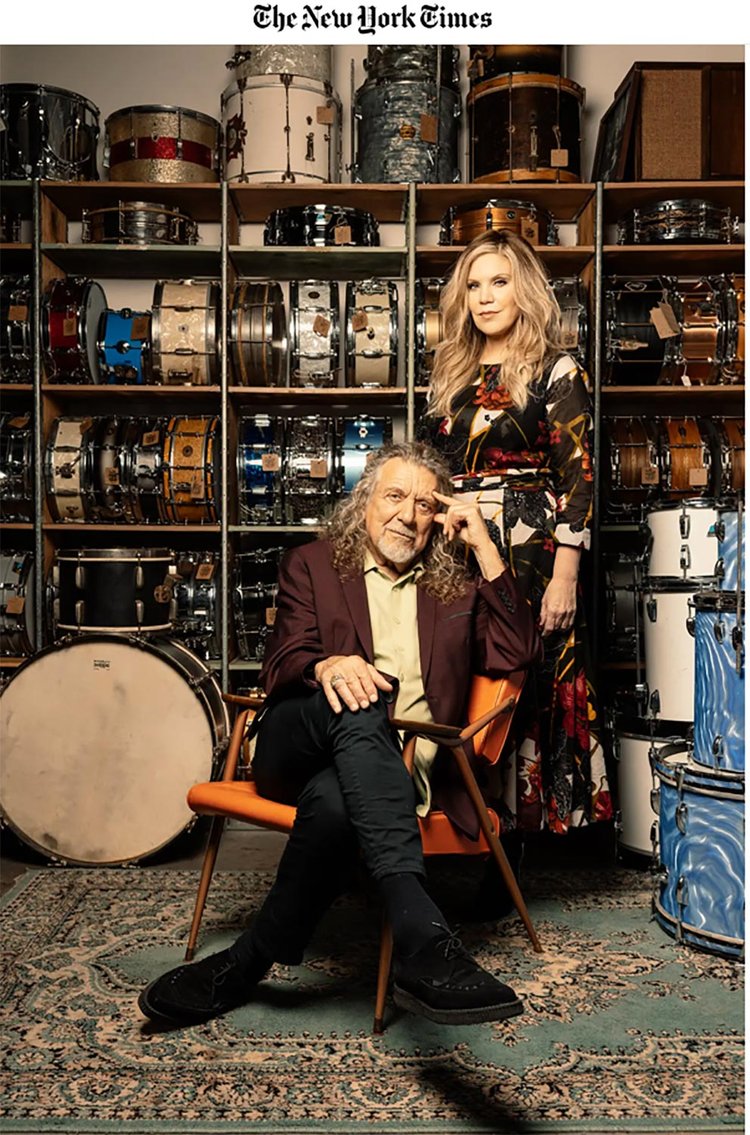 Portrait of a Robert Plant seated and a Alison Krauss standing in front of a wall filled with vintage snare drums, styled in elegant clothing with artistic flair, blending musical heritage and fashion in a rich, editorial composition featured by The New York Times.