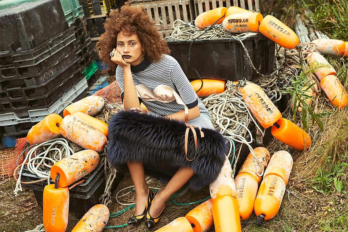 Fashion model seated among a pile of orange fishing buoys and tangled rope, wearing a textured dress with a satin accent, bold heels, and a luxurious dark fur stole, styled with striking handmade jewelry in a rugged coastal setting.
