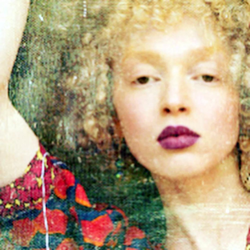 Close-up portrait of a model with curly blonde hair and bold plum lipstick, wearing a patterned red and blue garment and a large circular earring, seen through textured glass for a dreamy, vintage-inspired effect.