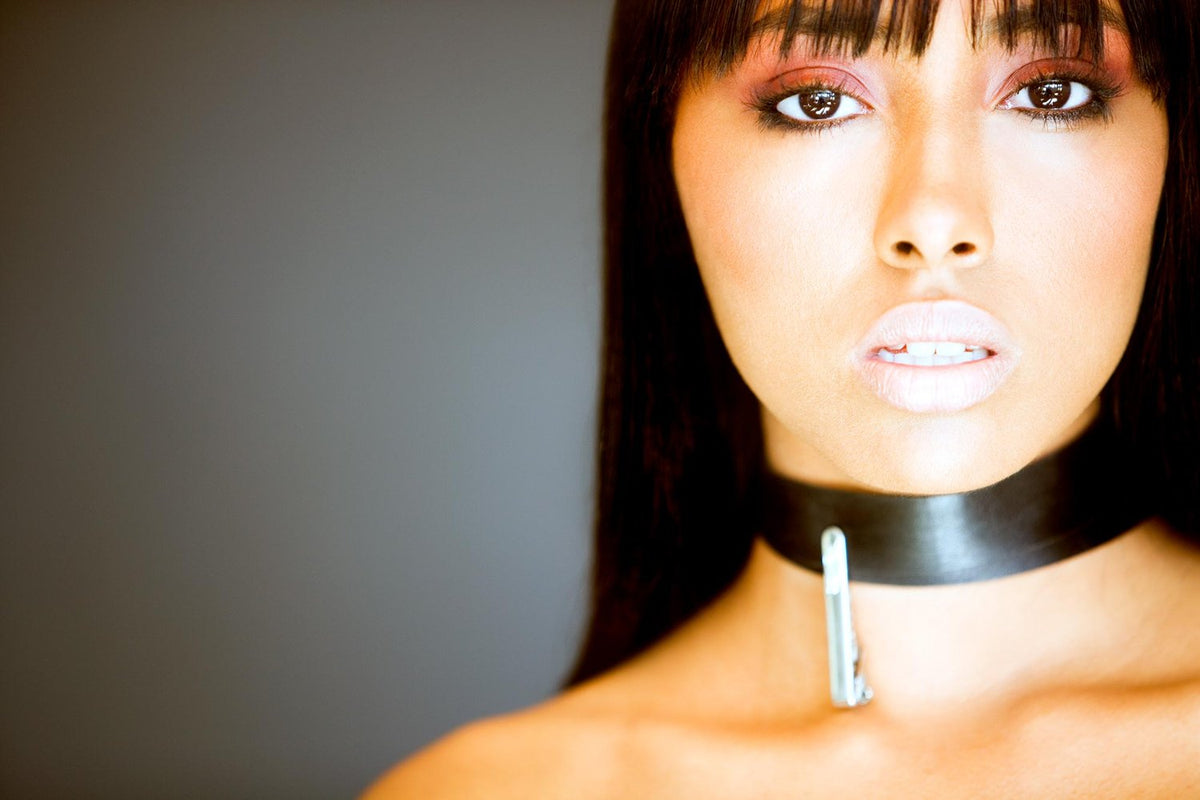 Close-up portrait of a woman with straight dark hair, wearing a bold black leather choker with a silver clasp, and soft-toned makeup featuring peachy lips and copper eyeshadow, set against a smooth, neutral background.