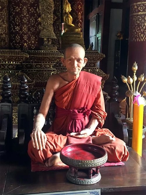 Photograph of a serene monk figure seated cross-legged in a richly ornate temple. Dressed in traditional saffron robes, the monk sits with a meditative posture, surrounded by dark wood carvings and golden statues in the background, evoking a sense of stillness and spiritual depth.