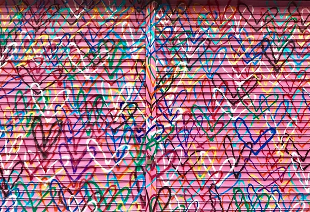 Image of a pink metal roll-down gate covered in hundreds of hand-drawn, multicolored hearts. The overlapping heart outlines create a dense, energetic pattern filled with vibrant colors like blue, red, yellow, green, and purple, giving the piece a joyful, chaotic feel—like street art love confetti.