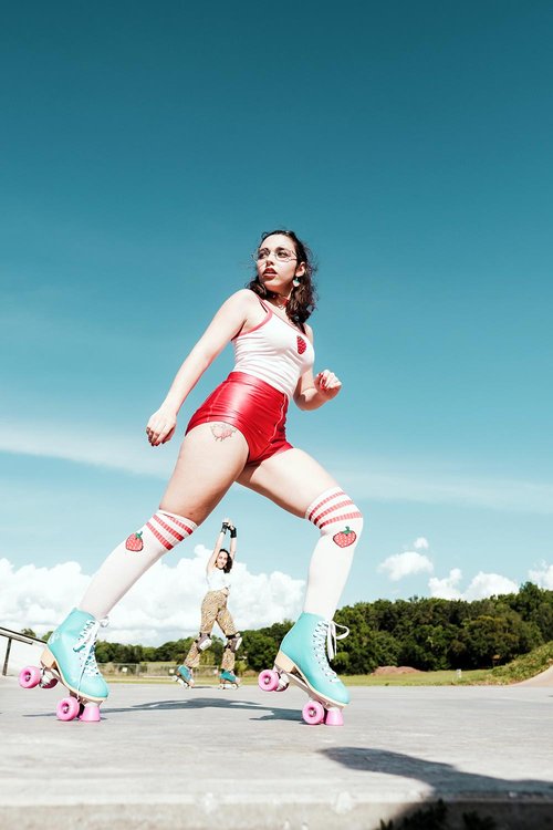 A confident woman in retro-style roller skates, red shorts, and a white tank top poses powerfully at a skate park under a clear blue sky, with another skater mid-jump in the background.