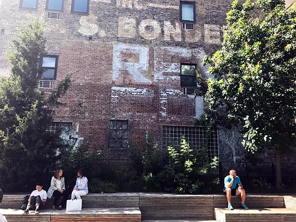 People sitting on wooden benches in front of a brick building with faded painted advertisements and greenery, under a sunny sky.