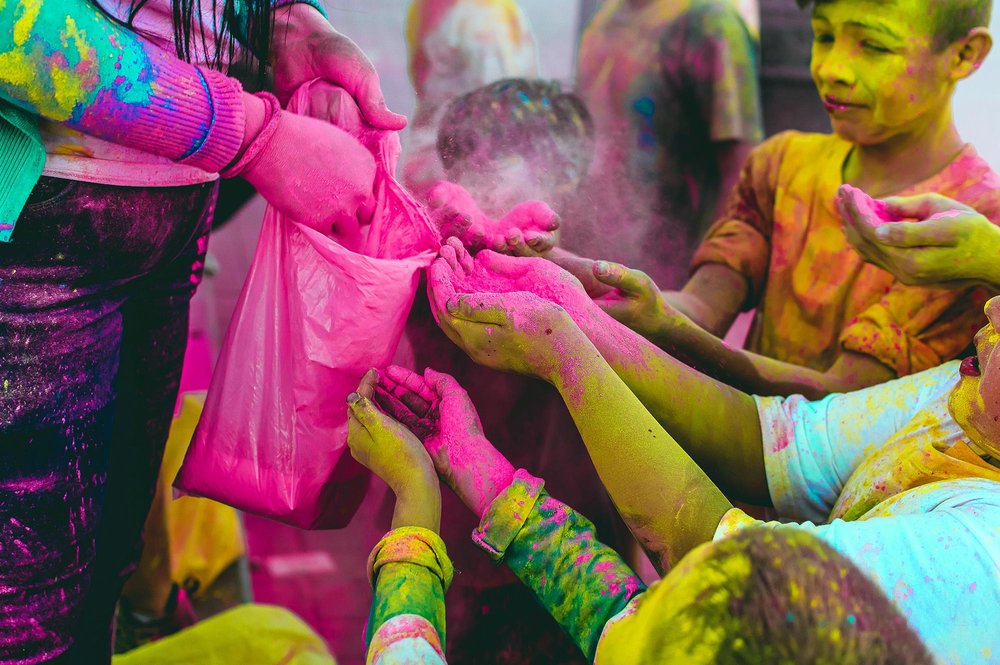 Children reaching out with brightly colored hands as pink powder is distributed from a bag, capturing a lively, festive moment reminiscent of Holi celebrations.