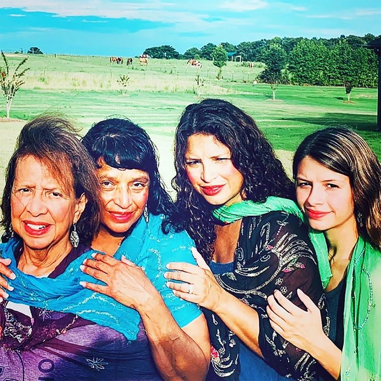 Four women from multiple generations stand closely together, smiling and embracing, with scarves in shades of turquoise and green draped over their shoulders. They are outdoors in a sunlit field with horses grazing in the background, evoking themes of family, heritage, and connection to nature.