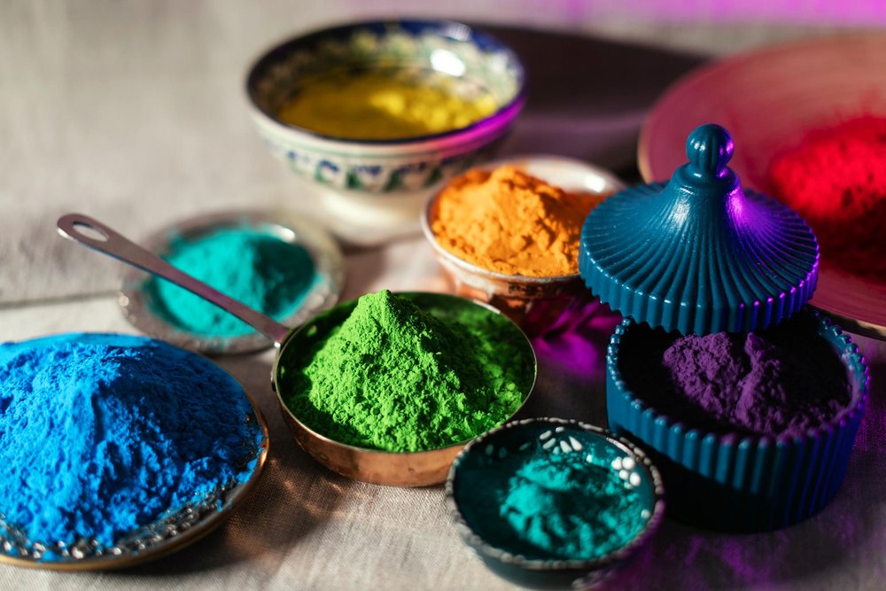 Colorful bowls and dishes filled with vibrant powdered pigments in shades of blue, green, orange, yellow, red, teal, and purple, arranged artfully on a light surface with dramatic lighting.