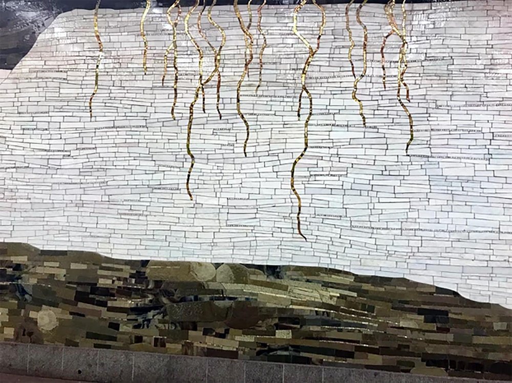 Wall mosaic featuring wavy vertical golden lines descending from the top over a white brick-like pattern, with an abstract earthy-toned base at the bottom.
