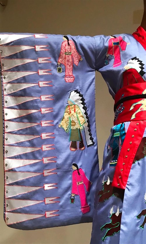 Close-up of a traditional garment with embroidered Indigenous figures and geometric patterns in vibrant colors on light purple fabric.