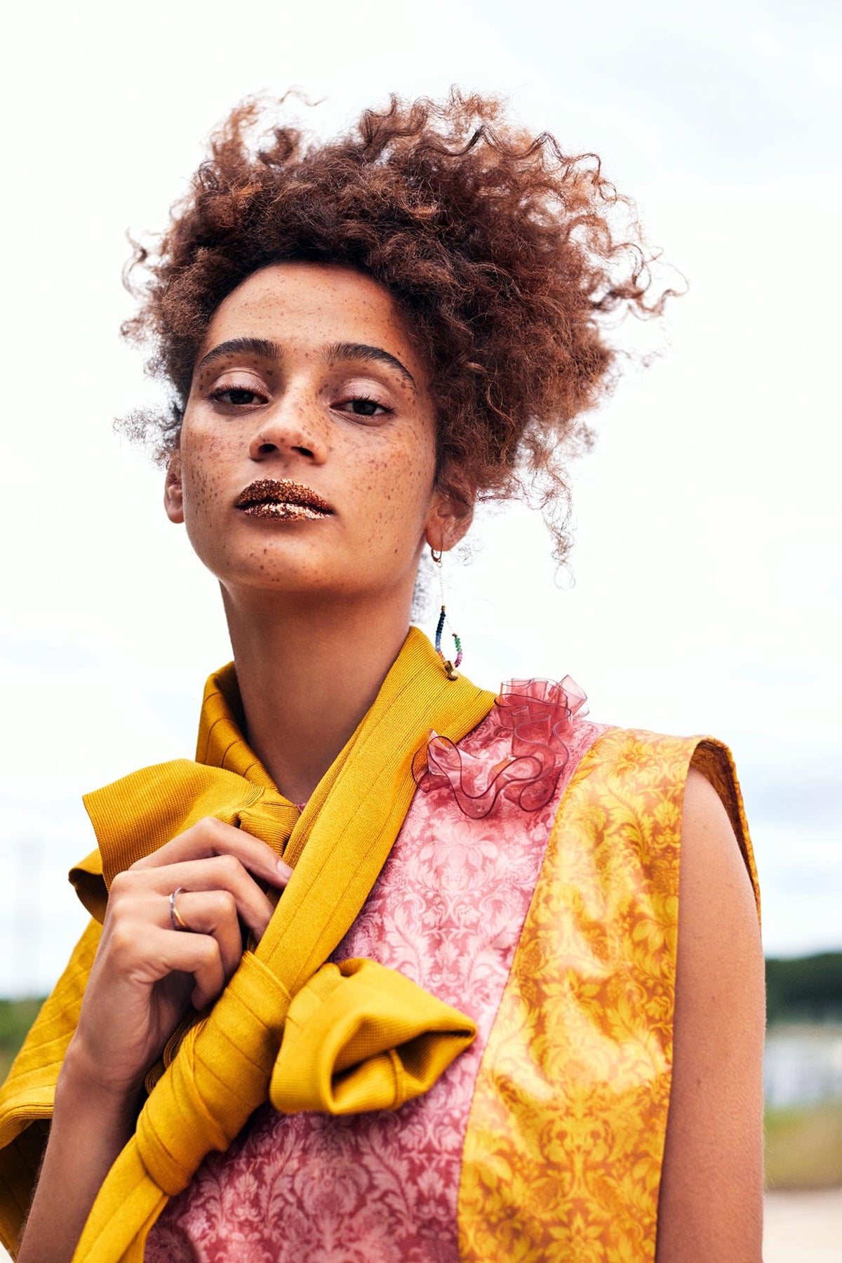Portrait of a woman with natural curly hair and gold glitter lips, wearing a vibrant outfit with mustard yellow and pink brocade fabric, looking confidently into the camera against a soft, cloudy sky.
