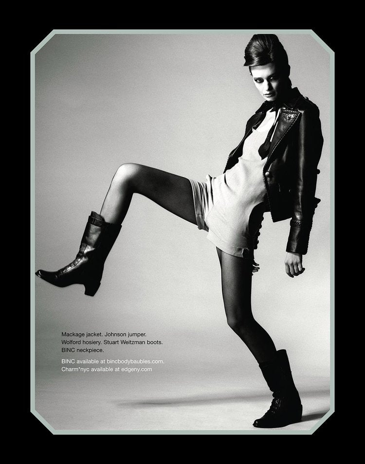 Black and white editorial shot of a model mid-step in a dynamic pose, wearing a leather jacket, mini jumper, fishnet tights, and black boots, styled with a statement handmade necklace that adds contrast and edge to the bold, minimalist look.