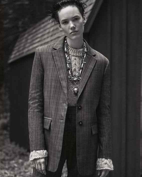 Black and white portrait of a model standing outdoors in a checkered suit layered over a textured sweater, styled with multiple chunky handmade necklaces that add earthy contrast and artisan detail to the structured, androgynous look.