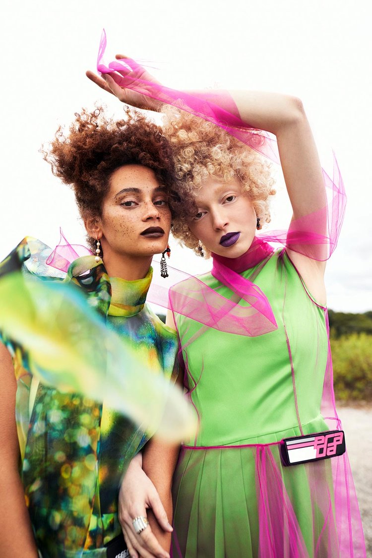 Two models standing close in bold, futuristic outfits—one in a shimmering multicolor dress and the other in neon green with pink tulle accents—wearing dramatic lipstick and statement handmade earrings, blending avant-garde fashion with natural surroundings.