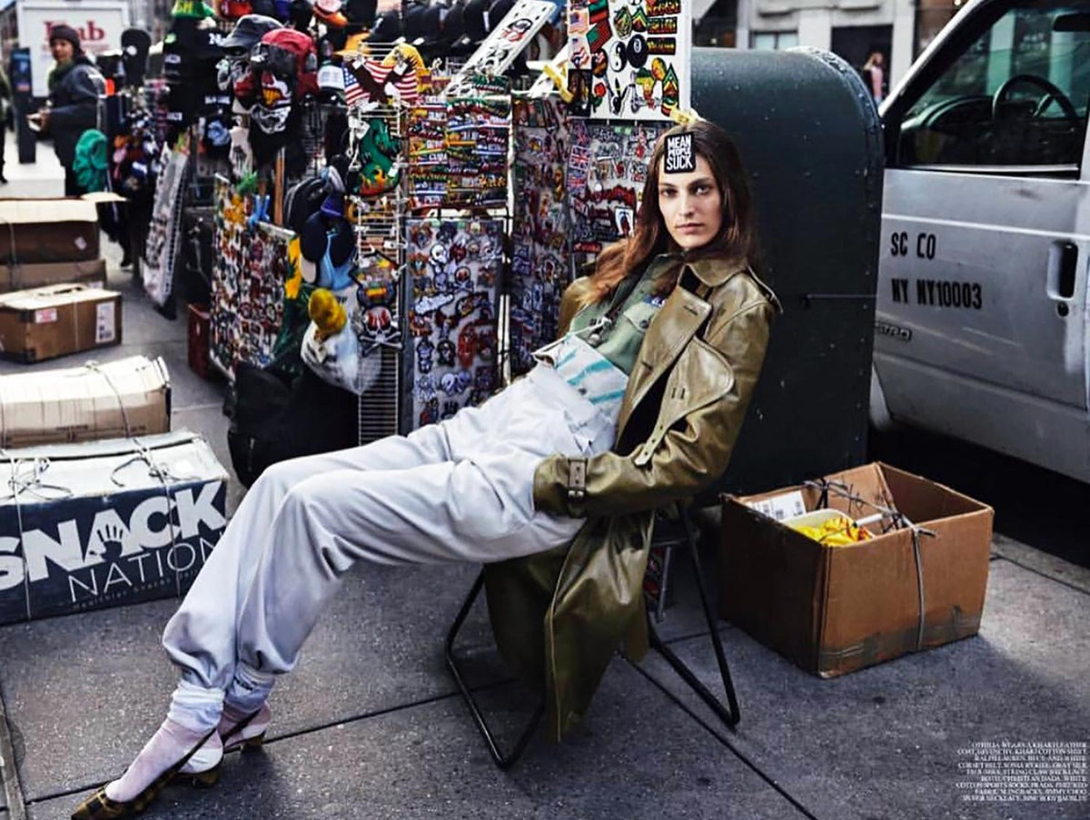 Model lounging in a street vendor scene wearing a long olive-green leather trench coat over a layered outfit with slouchy white pants and kitten heels, accented by bold jewelry and a “Mean People Suck” sticker on her forehead, blending high fashion with gritty urban surroundings.