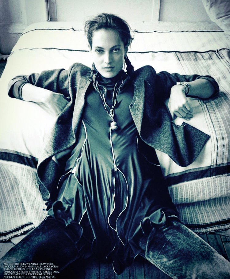 Fashion model reclining on a bed in a moody, monochromatic setting, wearing a long ruched dress layered with a wool blazer, textured boots, and striking handcrafted jewelry including long layered necklaces and bold silver earrings.